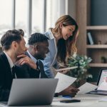 Three smiling business people analyzing charts and using laptop while working in the office together