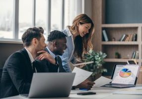 Three smiling business people analyzing charts and using laptop while working in the office together