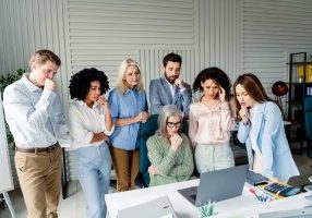 Colleagues gather around a laptop in an office, showing concentration and teamwork during a collaborative business session, illustrating professionalism and workplace collaboration.